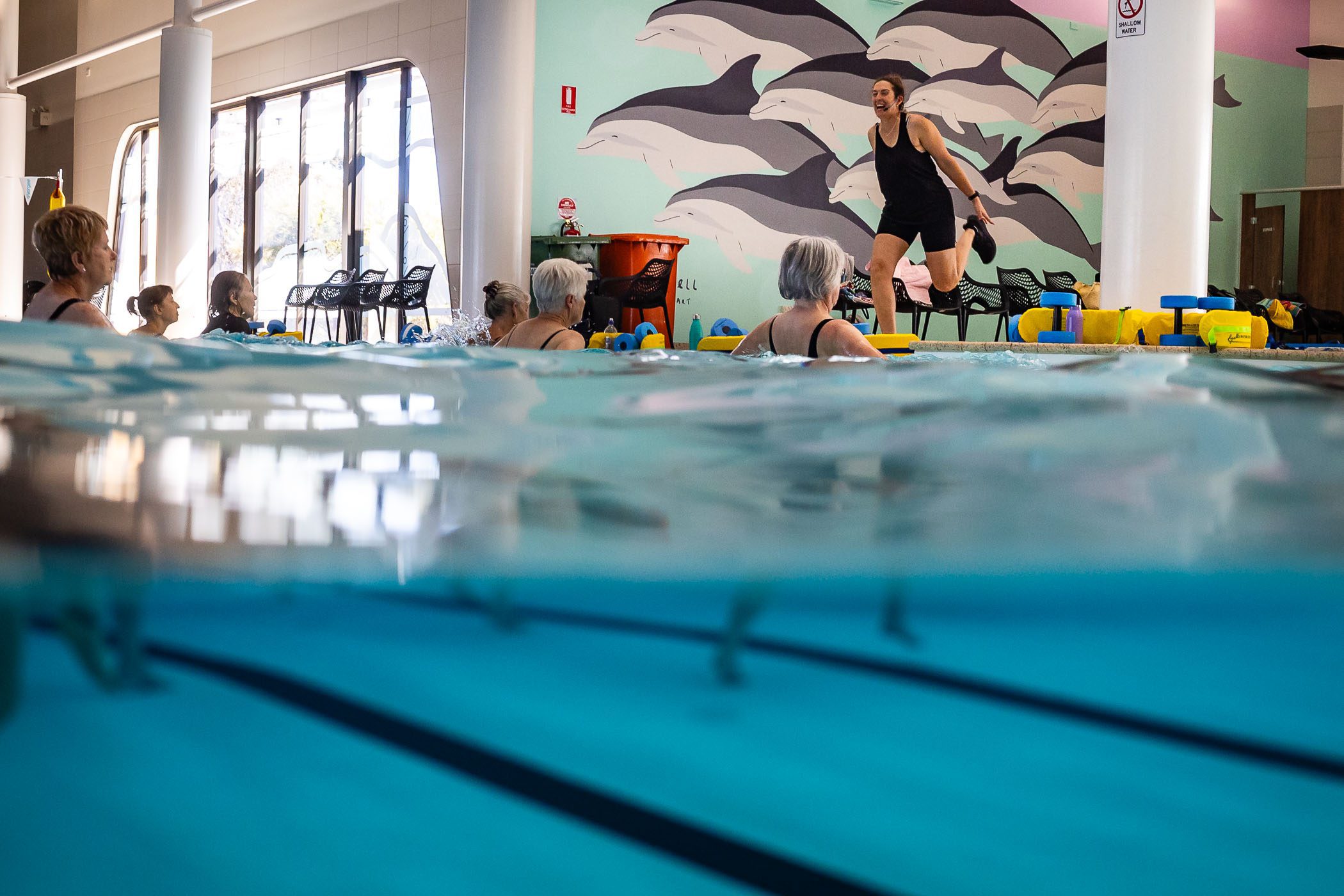 An aqua fitness class with participants in the water and the class instructor on the swimming pool side.