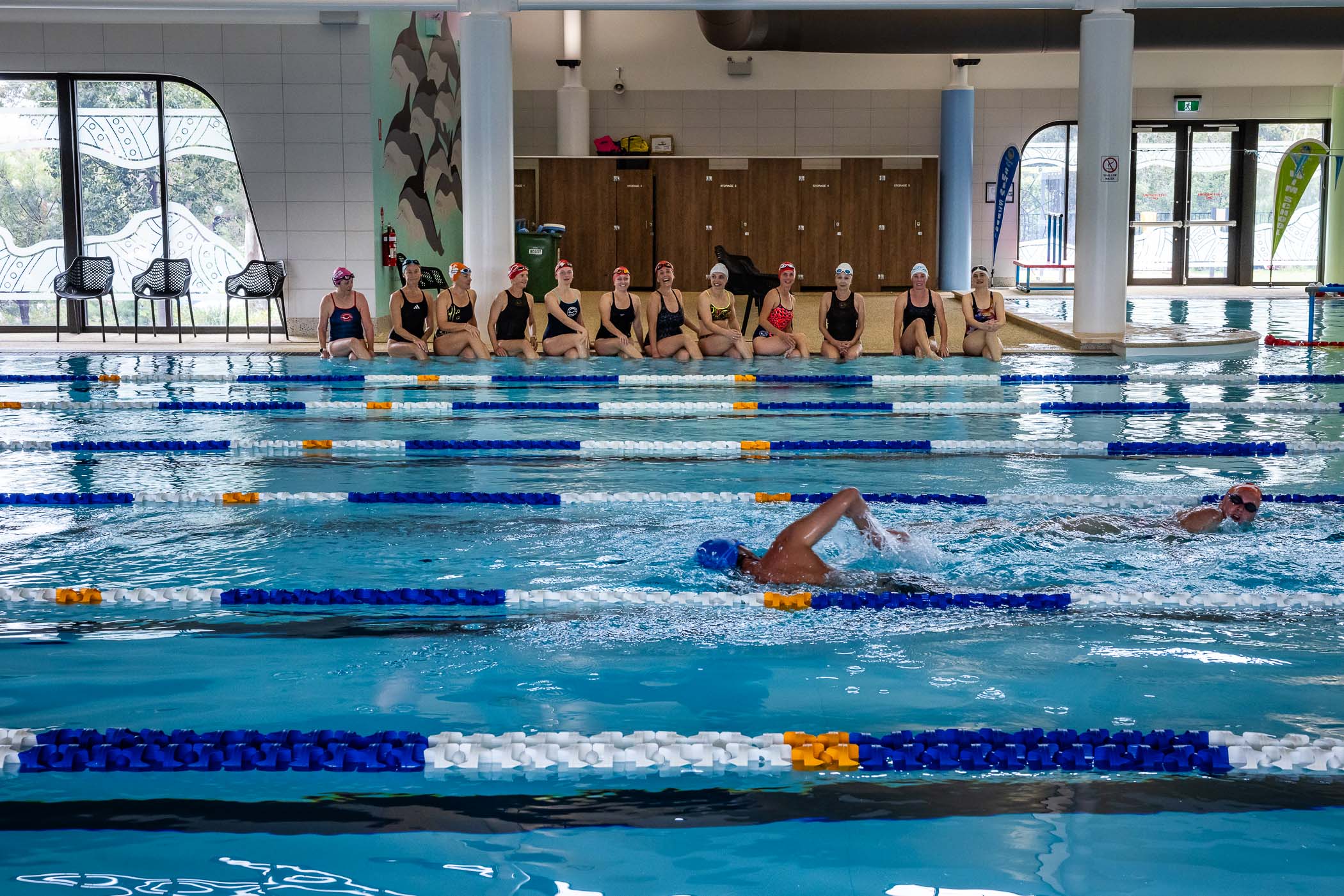 The Swimming Women club sits on the edge of Margaret River swimming pool. There are two swimmers doing laps in the water.