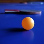 Vibrant orange ping pong ball with a table tennis racket on a blue table, emphasizing sport and leisure.